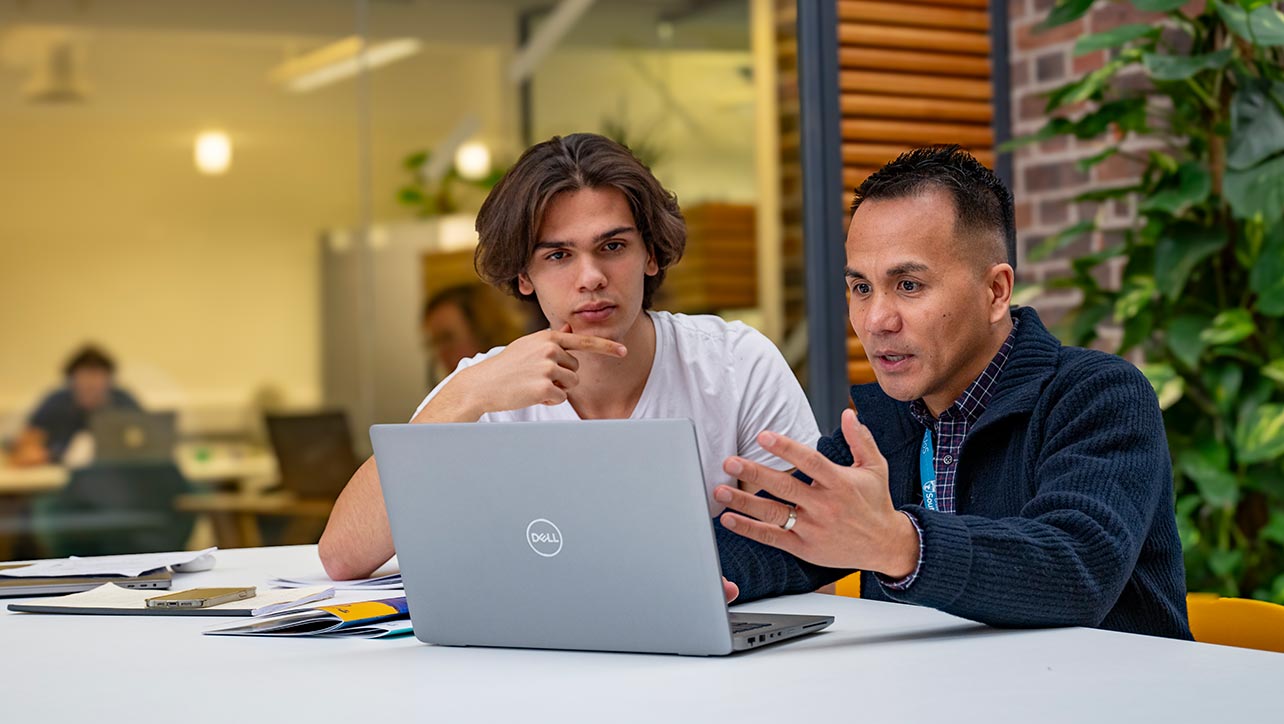 Two students sat next to each other, looking at the same laptop screen and having a discussion.