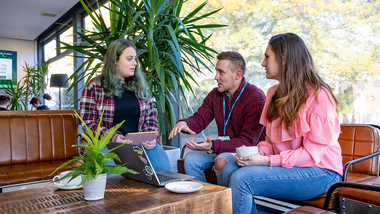 Three students are sat having a discussion with each other around a laptop sitting on a table. The student on the left is holding a tablet and the student on the right is holding a mug.