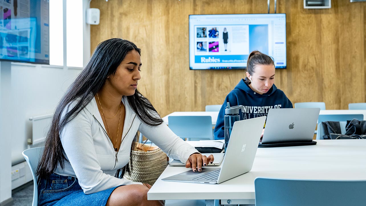 Two students are sat at a desk, each engrossed in their laptop screens. 