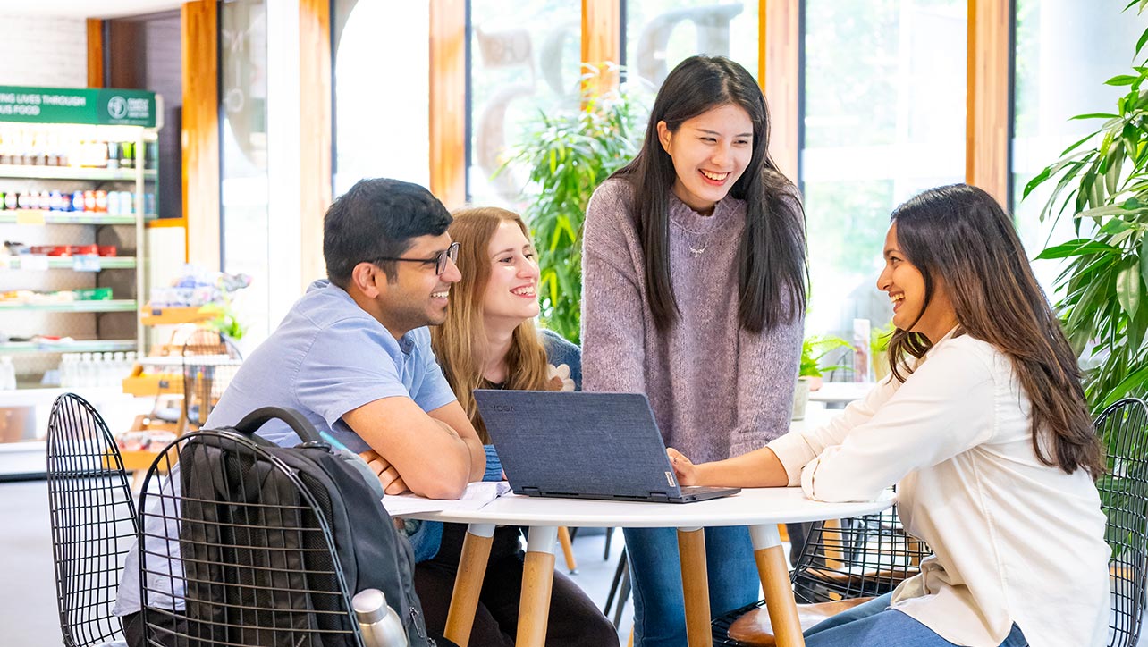 A group of four students, surrounding a table with a laptop and smiling at each other. One of them is standing. 