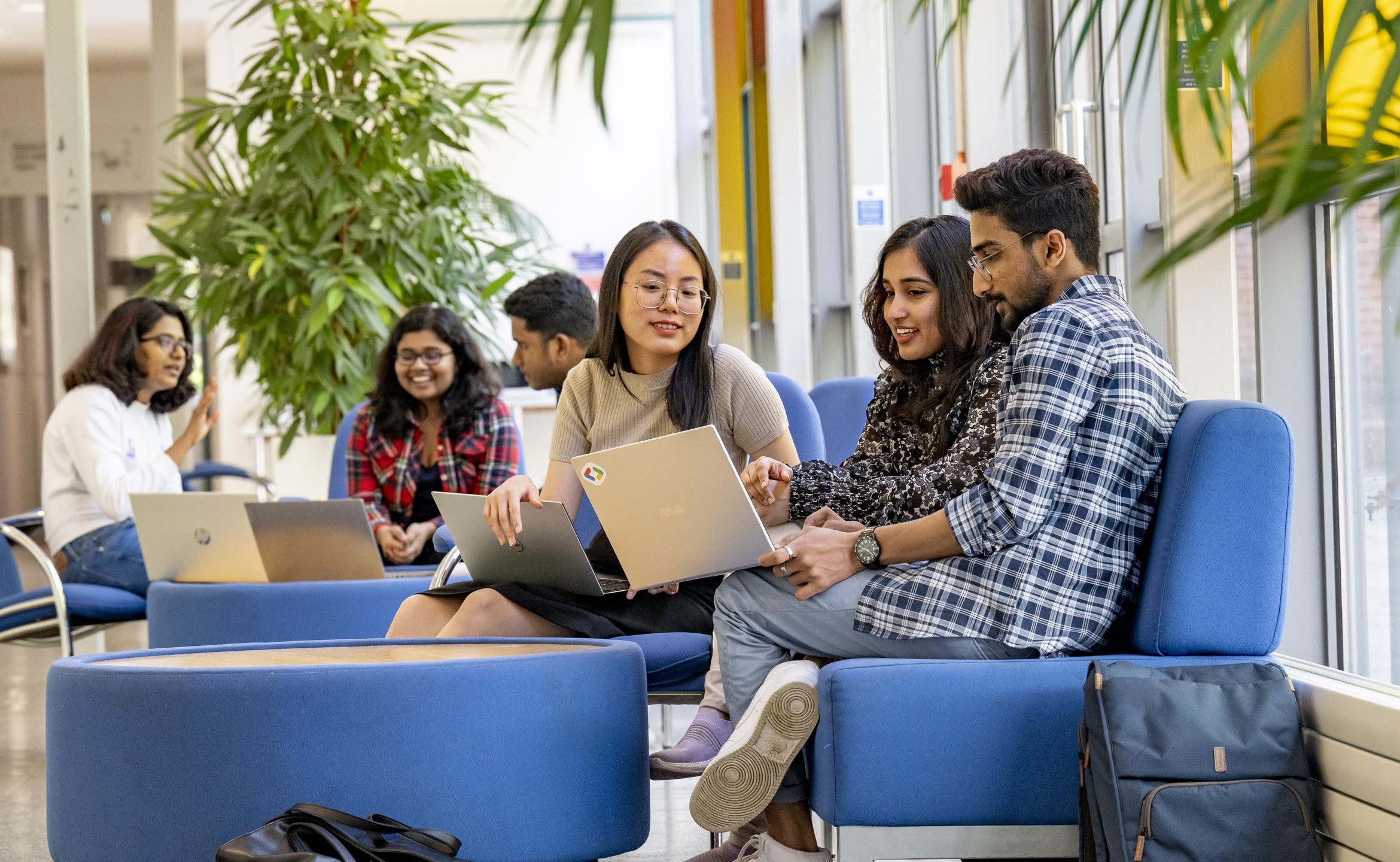 Three students sat in the foreground, looking at a laptop. There is also a group of three students in the background, chatting with each other.
