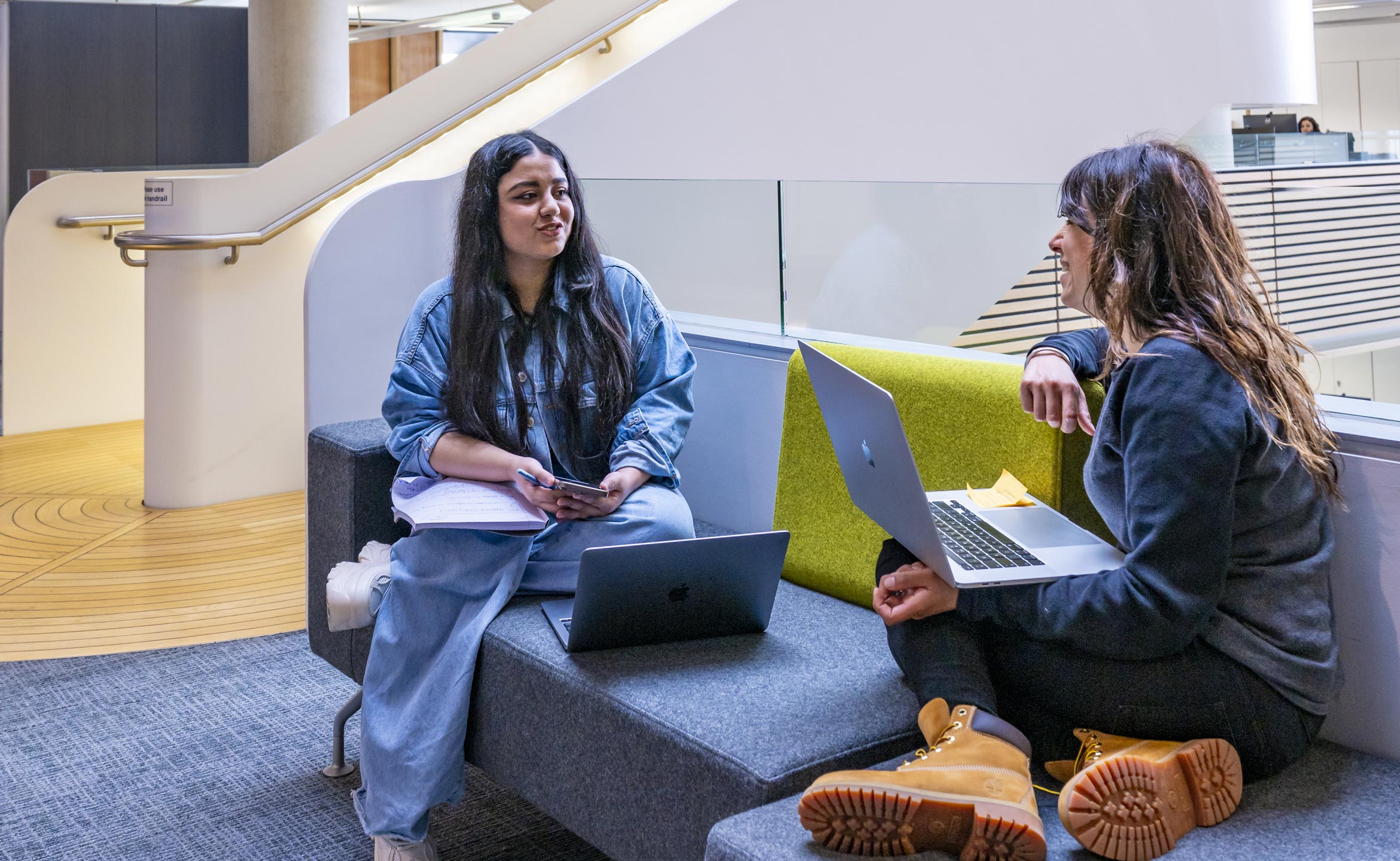 Two students sat in a university common area, facing each other at an angle and having a discussion. They each have a laptop.