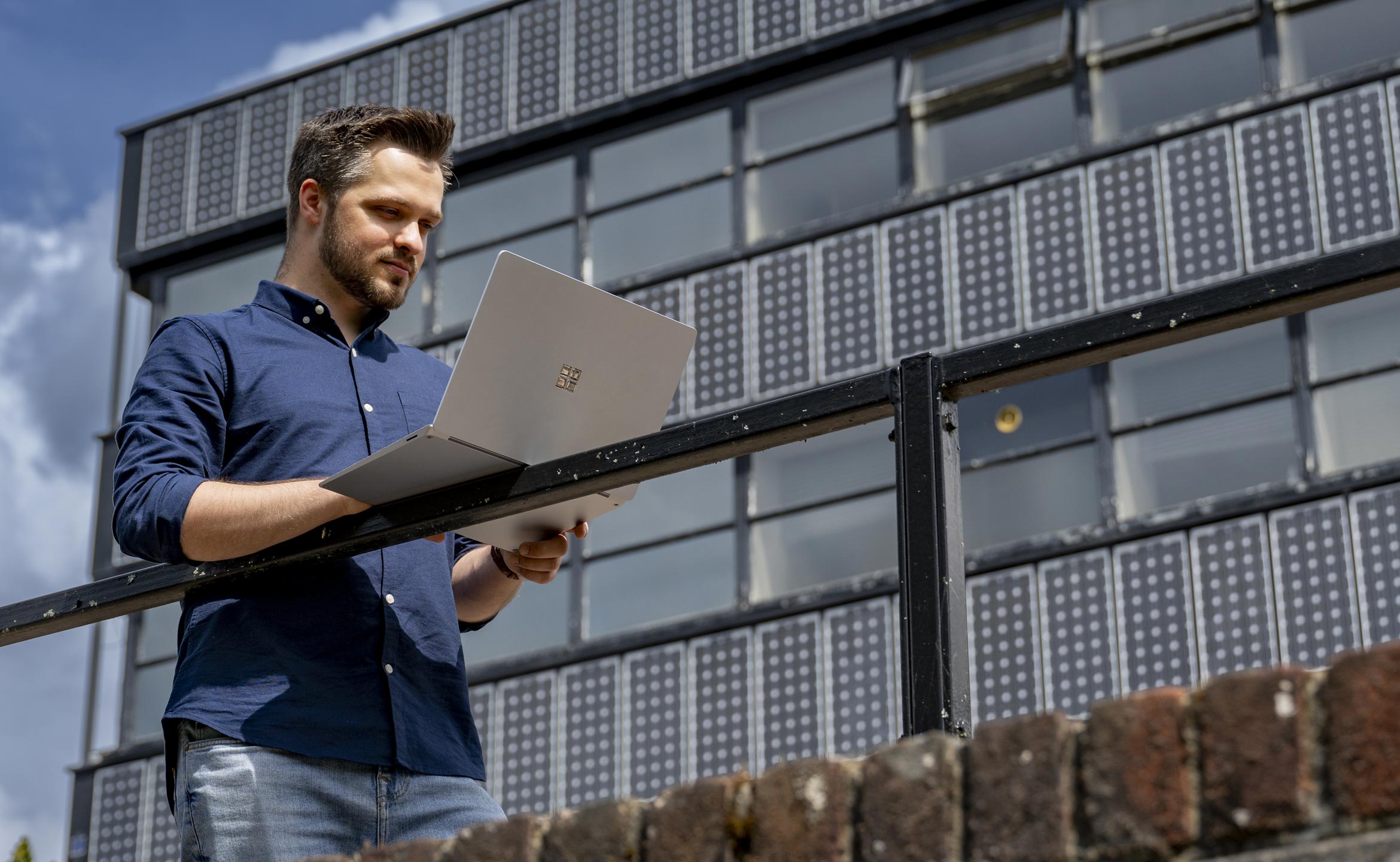 A student stood outside a university building, resting his laptop on a railing and looking at it.
