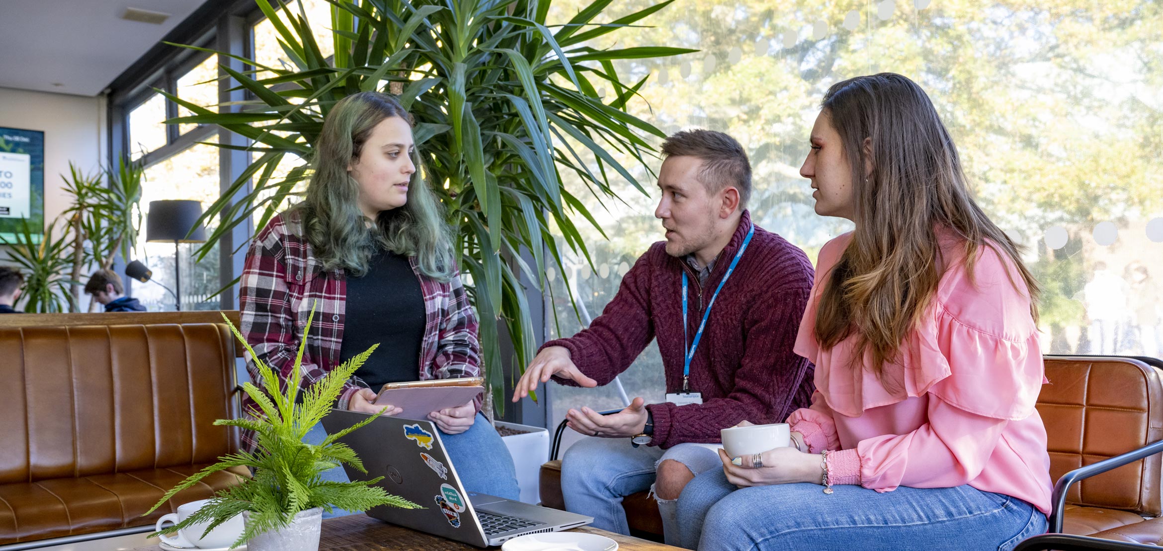 Three students are sat having a discussion with each other around a laptop sitting on a table. The student on the left is holding a tablet and the student on the right is holding a mug.
