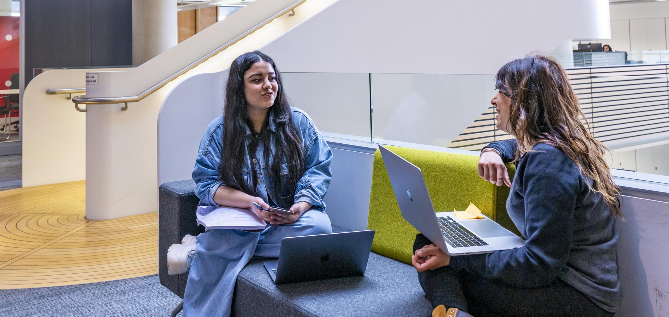 Two students sat in a university common area, facing each other at an angle and having a discussion. They each have a laptop.