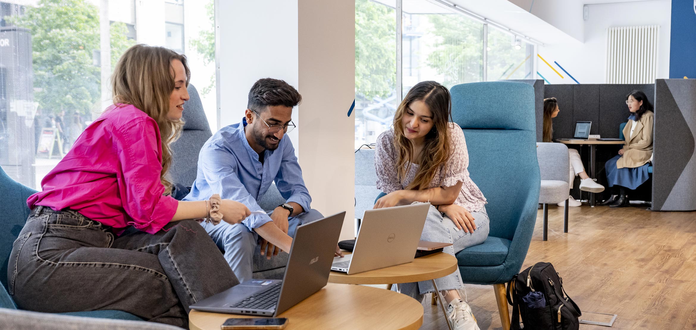 Three students are sat in a common area, looking at laptop screens inquisitively. 