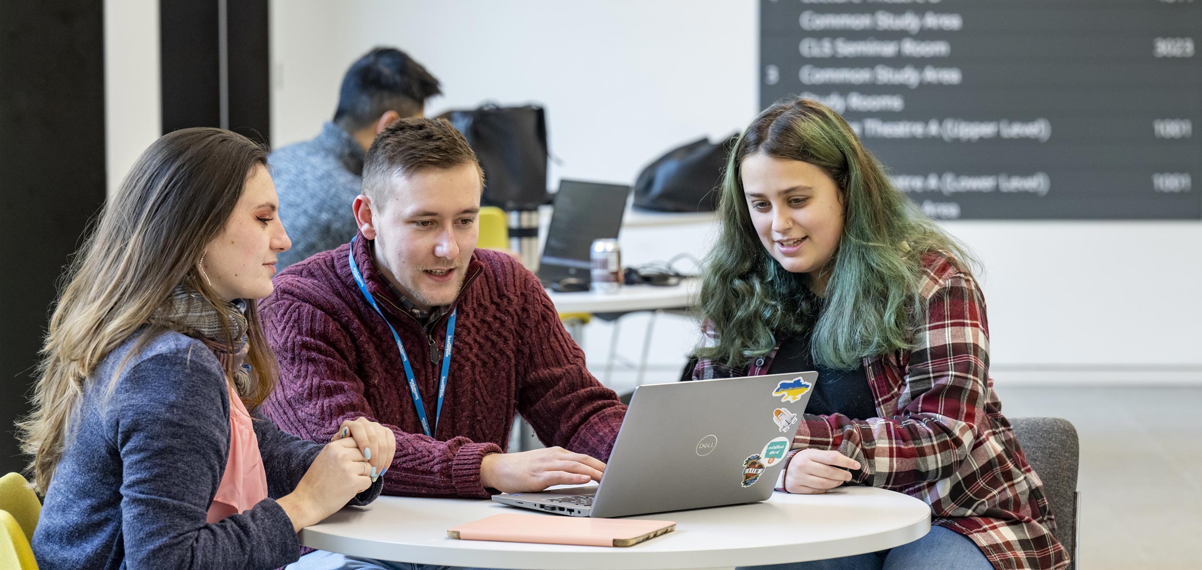 A group of three students all sat down at a round table, looking at a laptop screen.