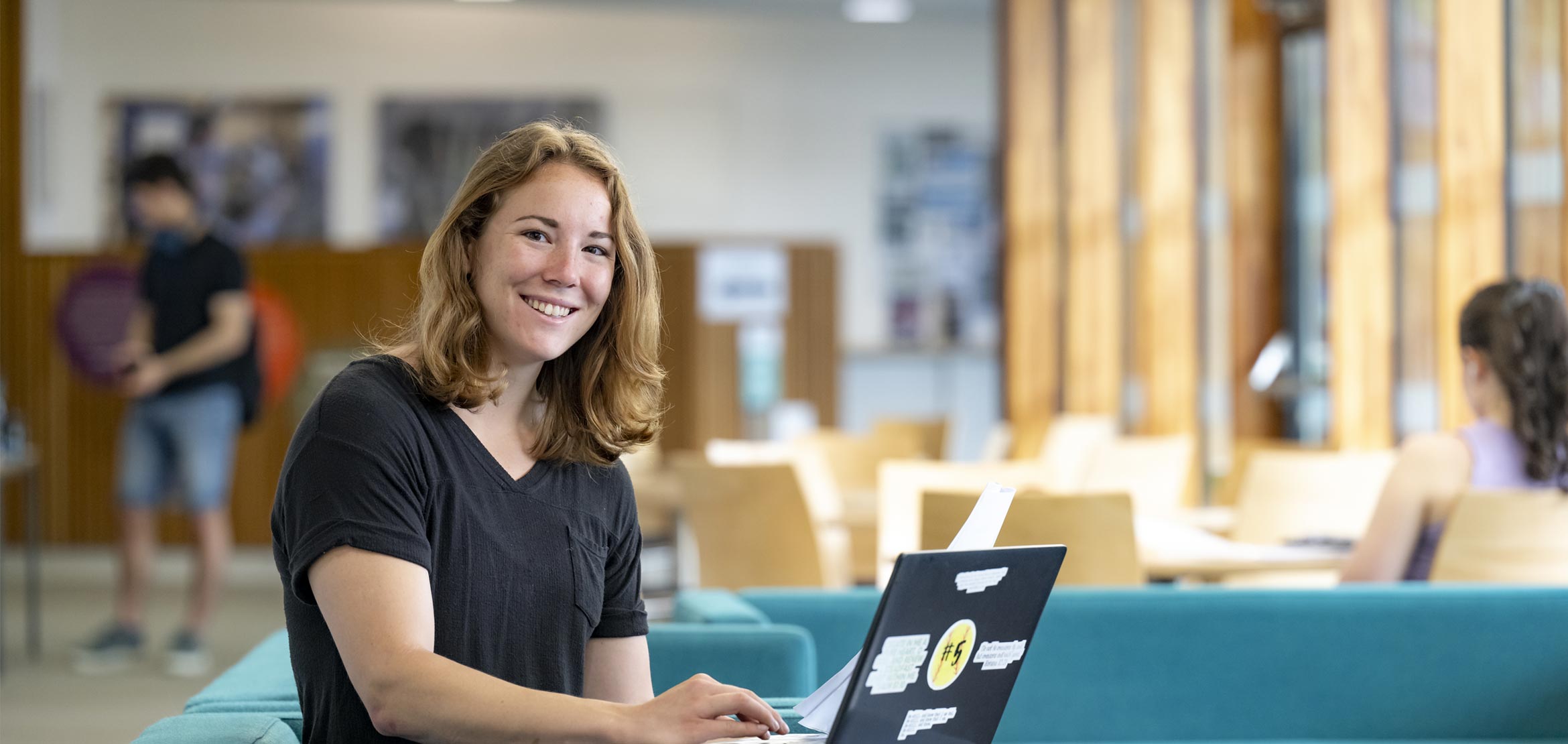 A student smiling to camera, sat down and working on a laptop.
